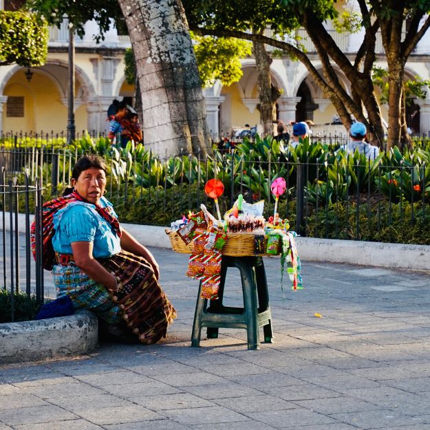 Woman in traditional clothing sitting on a street corner with a small cart displaying colorful items.