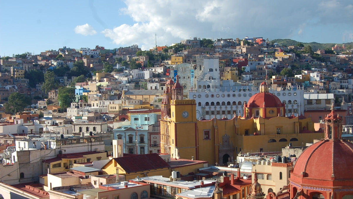 A scenic panoramic view of Mexico City 
with dense buildings, historic church towers, and red domes under a blue sky with white clouds.